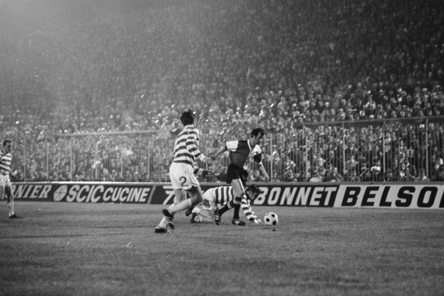Eine Gruppe von Männern, die auf einem Feld Fußball spielen, mit Zuschauern auf den Tribünen und einer Fahne mit der Aufschrift "Celtic v West Bromwich Albion - FA Cup Third Round" in Schwarz-Weiß.