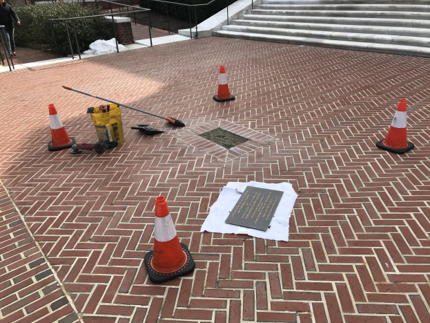 Backsteinweg mit Verkehrskegeln, einem Schild und einer Person auf der linken Seite, umgeben von Pflanzen, Geländern, Stufen und Gegenständen einschließlich eines Papiers mit Text, mit Bäumen und einem Gebäude im Hintergrund.