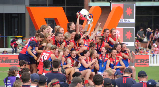 Eine Gruppe von Frauen-AFL-Spielerinnen in Sportuniformen feiert mit einem Pokal vor einem Publikum, mit Schildern, Geländern, Menschen und einem Gebäude mit Glasfenstern im Hintergrund und Gras am unteren Bildrand.