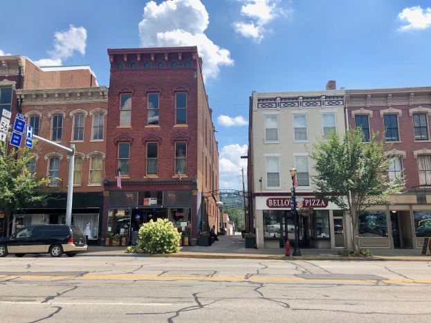 Eine Stadtstraße mit parkenden Autos, Gebäuden, Bäumen, Laternen, Schildern und einem Feuerhydranten unter einem bewölkten Himmel, die ein kommerzielles Grundstück zeigt.