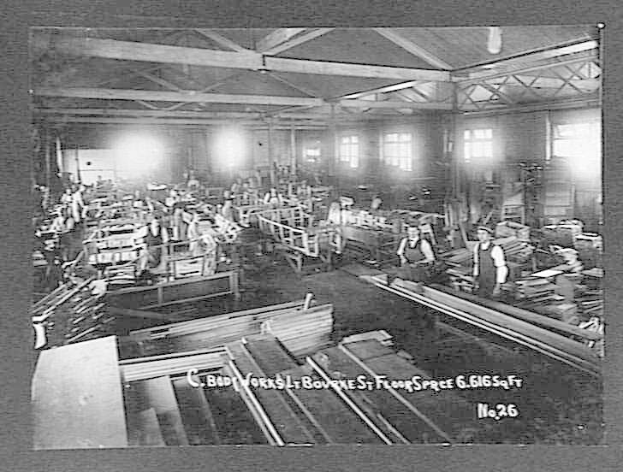 Schwarz-weißes Foto von Arbeitern in einer Fabrik, die hölzerne Gegenstände zusammenbauen, mit Lampen und Fenstern im Hintergrund und Text 'C. Body Works, St. Bourne's Floor Space, No. 26' unten.