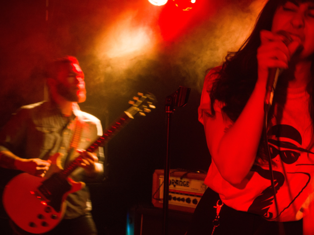 Eine Frau singt in ein Mikrofon auf der Bühne, während ein Mann eine akustische Gitarre im Hintergrund spielt, mit Deckenbeleuchtung, die die Bühne erhellt.