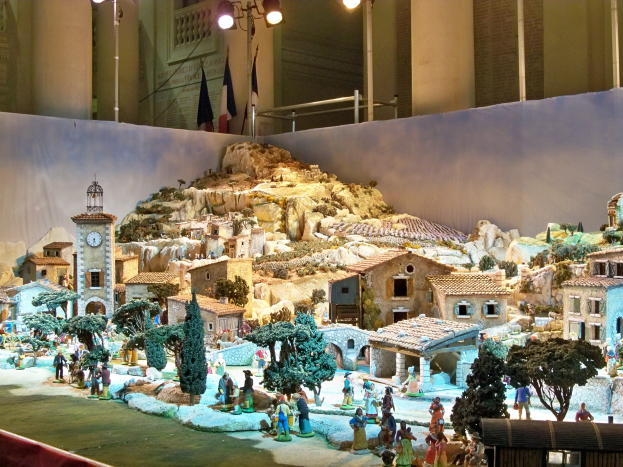 Ein detailliertes Miniatur-Modell einer Krippe in einem Museum ausgestellt, mit Figuren, Gebäuden, Bäumen, einem Kirchturm, Fahnen und beleuchtet vom Museumslicht.