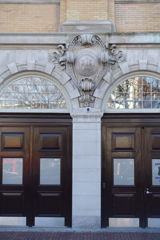 Zwei große hölzerne Türen mit Glasfenstern und Verzierungen an der Wand, erreichbar über einen Weg am unteren Bildrand.