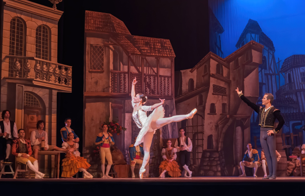 Zwei Tänzer performen auf einer Bühne im Royal Opera House in London, mit einem Publikum im Vordergrund, das auf Stühlen sitzt und steht, und einer Projektion von Gebäuden im Hintergrund.