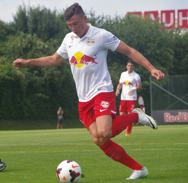Ein professioneller Fussballspieler tritt einen Ball auf einem grünen Feld mit Bäumen und einem klaren blauen Himmel im Hintergrund, wobei ein "RB Leipzig"-Schild sichtbar ist.