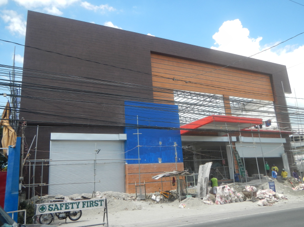 Baustelle mit einem "Sicherheit geht vor"-Schild am Gebäude, Fahrzeuge auf der Straße, eine Texttafel, Menschen in der Nähe, Bäume, Versorgungsmasten, Drähte und den Himmel im Hintergrund.