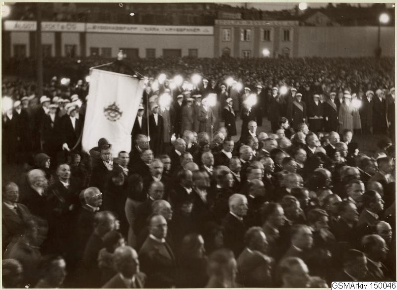 Schwarzes und weißes Bild einer großen Menge vor einem Gebäude, mit einer Person, die eine Fahne in der Mitte hält, Pfosten und Lichter im Hintergrund und Text unten, der "Gustav Klimts Rede an der Berliner Mauer" lautet.