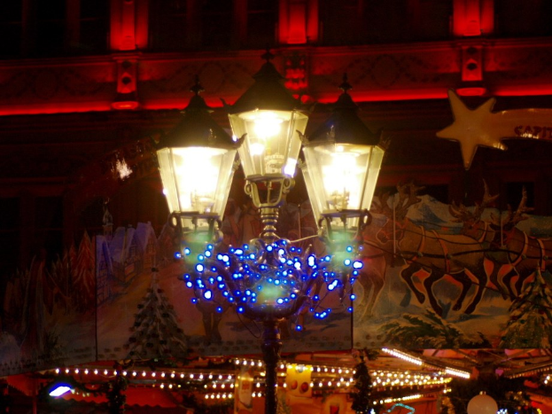 Eine Straßenlaterne in einer nächtlichen Stadt, geschmückt mit Lichtern und festlichen Gegenständen, steht vor einem Gebäude.