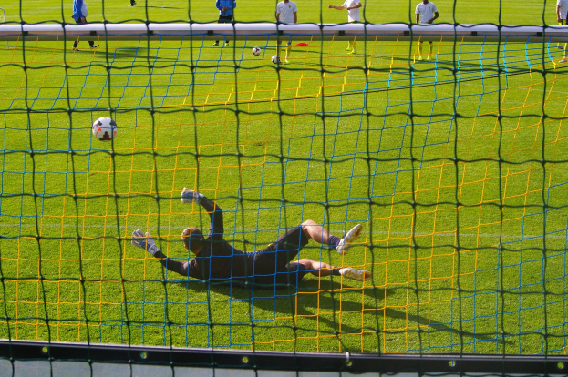 Ein Fußballtorhüter beim Abwehrversuch vor dem Tor, mit einem Netz im Vordergrund und einigen Personen im Hintergrund, während mehrere Fußballbälle auf dem Boden verstreut sind.