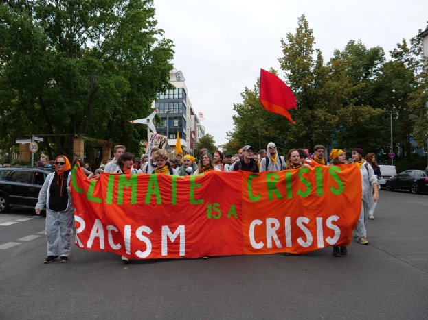 Eine Gruppe von Menschen marschiert eine baumbestandene Straße entlang und hölt ein Transparent mit der Aufschrift "Klima-Krise ist eine Krise", mit Gebäuden und Fahrzeugen im Hintergrund unter einem sichtbaren Himmel.