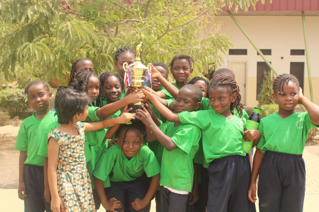 Gruppe von Kindern mit einem Pokal im Freien in einer Schulumgebung mit Bäumen, Pflanzen und einem Gebäude im Hintergrund.