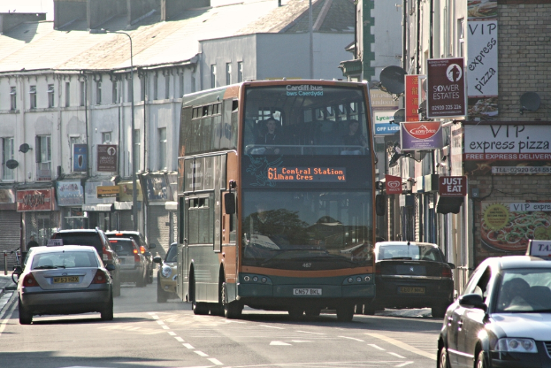 Eine Straße mit Autos und einem Bus im Vordergrund, mit Gebäuden mit Wänden, Fenstern, Tellern und Dächern im Hintergrund sowie Plakaten und Bannern an den Wänden und einem Pfahl mit einer Straßenlaterne.