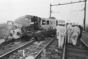 Ein Schwarz-Weiß-Bild einer Zugkollision mit einer Gruppe von Menschen in Helmen drumherum, Strommasten, Bäumen, Gebäuden und einem klaren Himmel im Hintergrund.