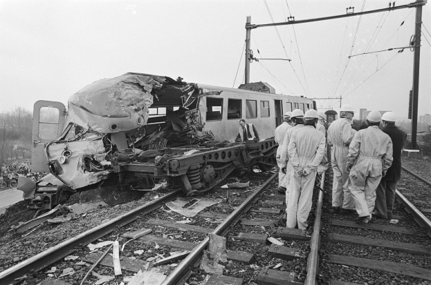 Ein Schwarz-Weiß-Bild einer Zugkollision mit einer Gruppe von Menschen in Helmen drumherum, Strommasten, Bäumen, Gebäuden und einem klaren Himmel im Hintergrund.