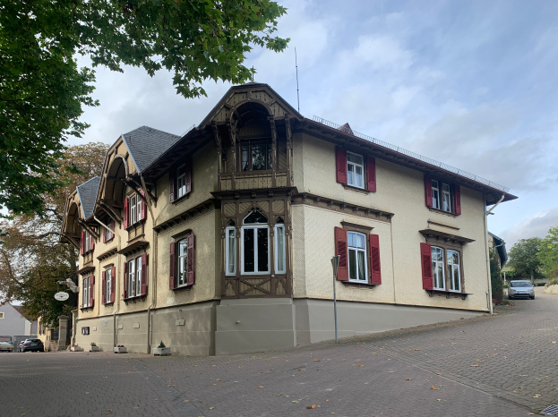 Ein großes Gebäude mit roten Fensterläden, umgeben von Bäumen und Fahrzeugen auf der Straße, unter einem bewölkten Himmel in Heidelberg, Deutschland, verfügbar zur Miete als Wohnung.
