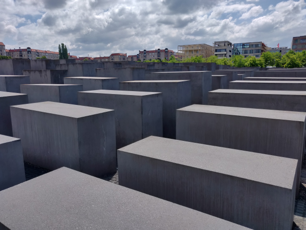 Konkreter Holocaust-Denkmal mit rechteckigen Blöcken, die in einem Gittermuster angeordnet sind und von Bäumen, Gebäuden und einer bewölkten Himmel umgeben sind.