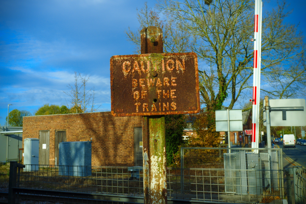 Warnschild an einer Bahnschiene neben einem Metallzaun, mit Bäumen, Strommasten, einem Gebäude, Containern, Fahrzeugen und einem bewölkten Himmel.