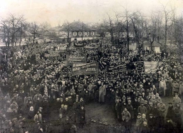 Eine große Menschenmenge mit Schildern steht vor einem Gebäude, mit Bäumen und Fenstern im Hintergrund, auf einem Schwarz-Weiß-Bild.