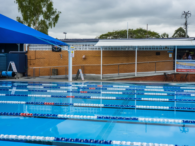 Großer Schwimmbad mit Bahnentrennern, umgeben von einem Zaun, einer Hütte, einem Banner, Stühlen, einem Tisch, einem Schild, Polen, einem Straßenmast, Drähten, Bäumen und einem bewölkten Himmel.