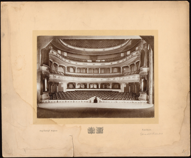 Schwarzes und weißes Foto des Auditoriums der Royal Albert Hall, das die Bühne, Reihen von Stühlen, Säulen, Geländer und Text mit einem Logo unten zeigt.