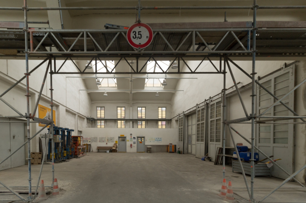 Ein gro\sser Lagerhalle mit ausgedehnten Ger"usten, Verkehrskegeln auf dem Boden, Metallstangen, ein Schild, verschiedene Maschinen, eine T"ur, Fenster, eine Wand und ein Dach mit Deckenleuchten.