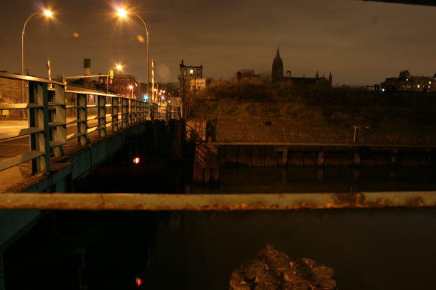 Eine Brücke überspannt einen Fluss mit mehreren Straßenlaternen entlang ihrer Länge und zahlreiche Gebäude sind im Hintergrund sichtbar.
