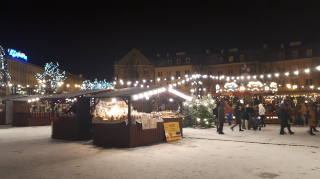 Lebendiger Weihnachtsmarkt in einer verschneiten Nacht mit Menschen, Buden, Pflanzen, Gebäuden, Schildern und einem bewölkten Himmel.