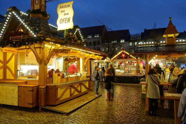 Ein nächtlicher Weihnachtsmarkt auf einer Kopfsteinpflasterstraße mit festlich beleuchteten Ständen, Menschen, die durch die Gänge schlendern, und Gebäuden mit beleuchteten Fenstern im Hintergrund.