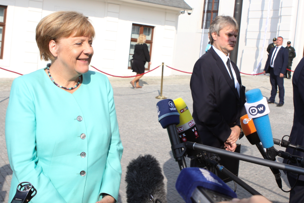 Bundeskanzlerin Angela Merkel spricht vor einem Gebäude mit Fenstern zu Reportern, umgeben von einer Gruppe von Menschen, einige halten Mikrofone mit Ständern, und ein Seil ist an einen Pfahl gebunden, unter einem bewölkten Himmel.
