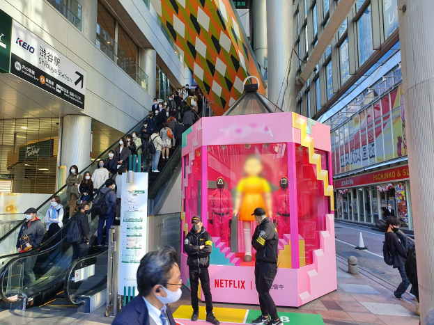 Eine Gruppe von Menschen steht vor einem Gebäude mit Glaswänden und Texttafeln, einige tragen Masken, während eine Rolltreppe mit Menschen zu sehen ist; eine rosafarbene Box in der Mitte zeigt Schaufensterpuppen und trägt den Text "Netflix's new Netflix booth in Tokyo."