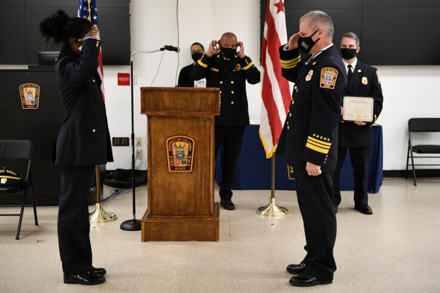 Gruppe uniformierter Personen in Masken auf einem Podium bei einer Vereidigung eines neuen Feuerwehrchefs, der eine Urkunde hält, mit Flaggen, Stühlen, Tischen und einer Wand im Hintergrund.