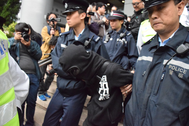 Eine Gruppe von Polizeibeamten steht vor einer Menge, einige halten Kameras, mit einem Gebäude mit Säulen und beleuchteter Decke im Hintergrund und Bäumen auf der linken Seite.