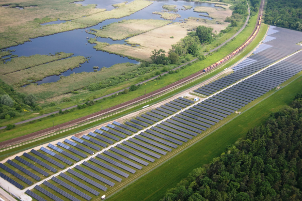 Luftbild einer Solarfarms mit Solarpanelen in einem Feld, umgeben von Bäumen, Gras, Pflanzen und Wasser, mit einem Zug auf einer nahen Bahnstrecke.