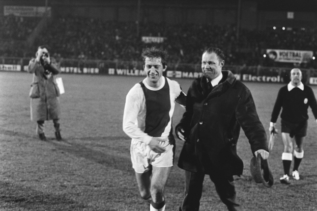 Ein Schwarz-Weiß-Foto einer Gruppe von Männern, die auf einem Fußballfeld gehen, mit Zuschauern auf den Tribünen und einem Mann in der Mitte mit fußfreundlicher Kleidung.