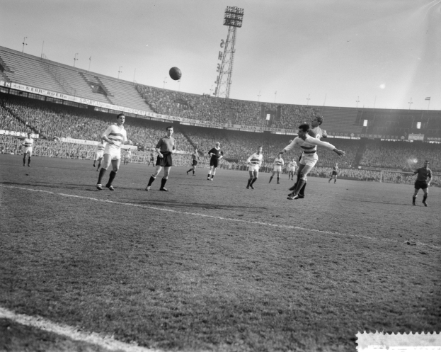 Eine Gruppe von Männern, die Fußball auf einem Feld spielen, mit einem Stadion, Flutlicht und einem klaren Himmel im Hintergrund.