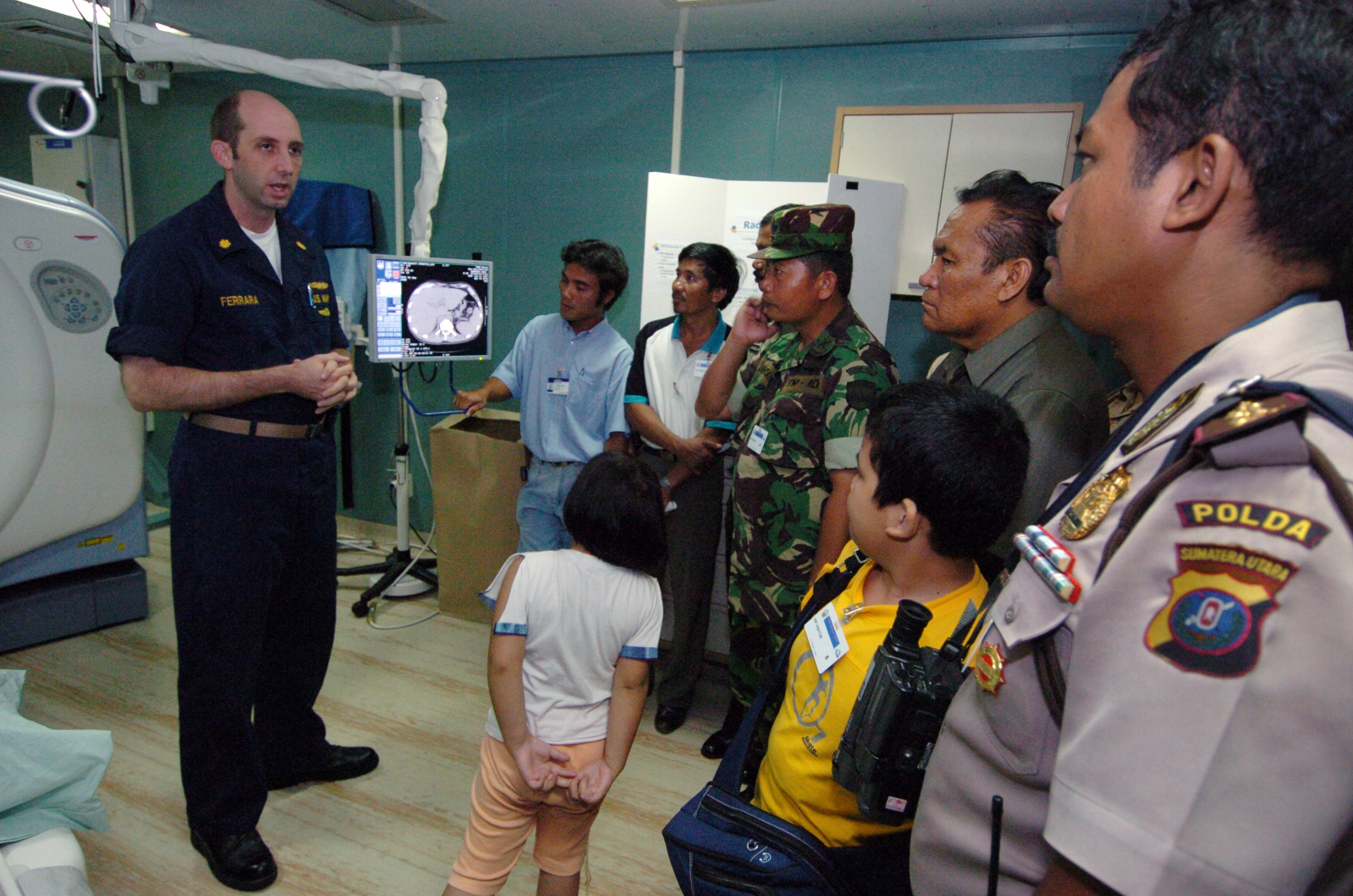 Gruppe von Menschen in einem Krankenhauszimmer mit einem Mann, der in ein Mikrofon spricht, einem drapierten Bett auf der linken Seite, medizinischen Geräten im Hintergrund, einem Röntgenmaschinenständer, einer Texttafel, einer Tür und Deckenleuchten.