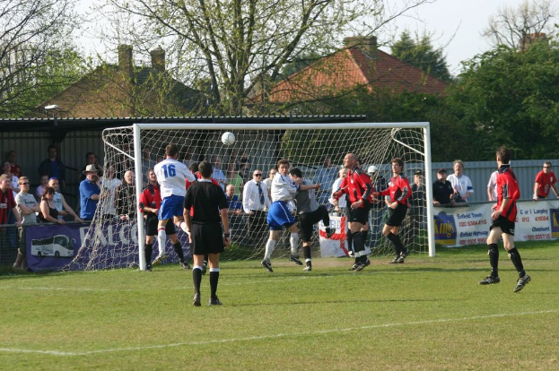 Fußballspieler bei einem Spiel auf dem Feld, mit einem Tor im Hintergrund und Zuschauern dahinter, während in der Ferne Bäume und Häuser sichtbar sind.