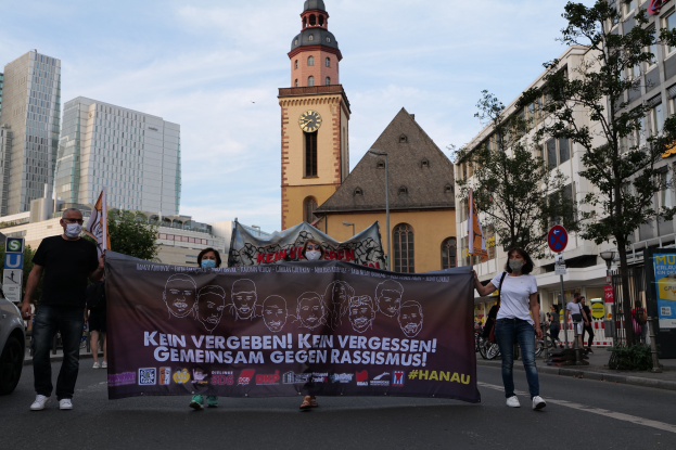 Eine Gruppe von Menschen mit Masken, die eine Straße entlanggehen und ein Banner halten, mit einem geparkten Auto links, Gebäuden, Bäumen, Schildern, Pfosten, einem Uhrenturm und einem klaren blauen Himmel im Hintergrund.