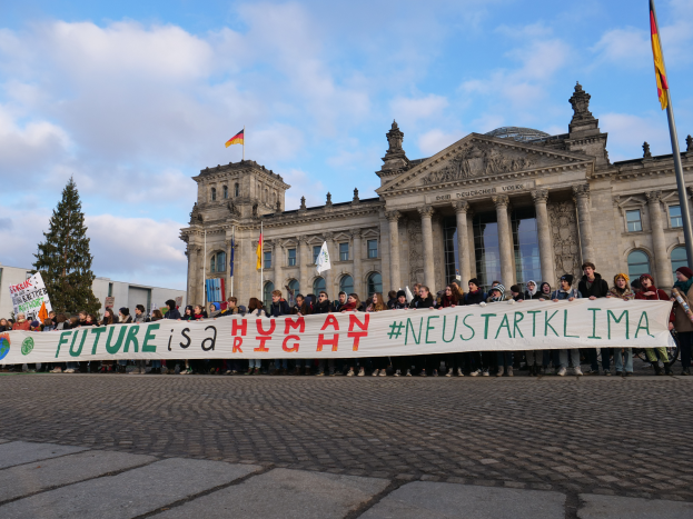 Eine Gruppe von Menschen steht vor dem Reichstag in Berlin und hält ein Transparent mit der Aufschrift "Zukunft ist ein Menschenrecht". Das Gebäude ist mit Säulen, Fenstern und Bögen geschmückt und von Bäumen und Fahnen umgeben.