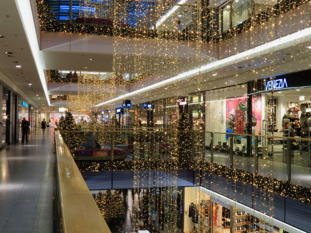 Ein festlich geschmücktes Einkaufszentrum mit Weihnachtsbeleuchtung, das Menschen zeigt, die durch die Gänge schlendern, Geschäfte mit Schildern und Glasgeländer sowie von der Decke hängende Lichter.