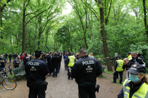 Eine Gruppe von Polizisten steht vor einer Menschenmenge, einige tragen M√∞tzen und Masken, mit Fahrr√°dern und einer Bank im Vordergrund und Bäumen und Himmel im Hintergrund, wahrscheinlich während einer Anti-Terror-Demonstration in Berlin.