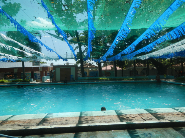 Großer Swimmingpool mit blauen und grünen Bändern, die von der Decke hängen, umgeben von Häusern, Bäumen und einem klaren blauen Himmel, mit Menschen, die das Wasser und die Sonne genießen.