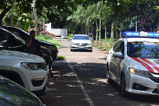 Ein Polizeiwagen steht auf der Seite einer Straße neben anderen Fahrzeugen, mit einer Person auf der linken Seite, die ein Mobiltelefon hält, und einem Hintergrund mit Bäumen, Schildern, Pfählen, Pflanzen, Gras und einer Wand.