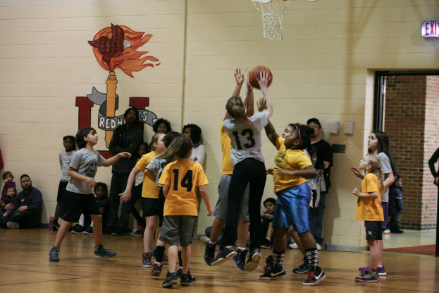 Kinder in gelben und grauen T-Shirts spielen Basketball, mit einem Mädchen in einem grauen T-Shirt mit der Nummer 13, das einen Wurf versucht. Im Hintergrund sitzt ein Mann in einem blauen Hemd auf dem Court mit einer weißen Wand mit Text dahinter und rechts daneben ein Ausgangsschild und ein rotes Backsteingebäude.