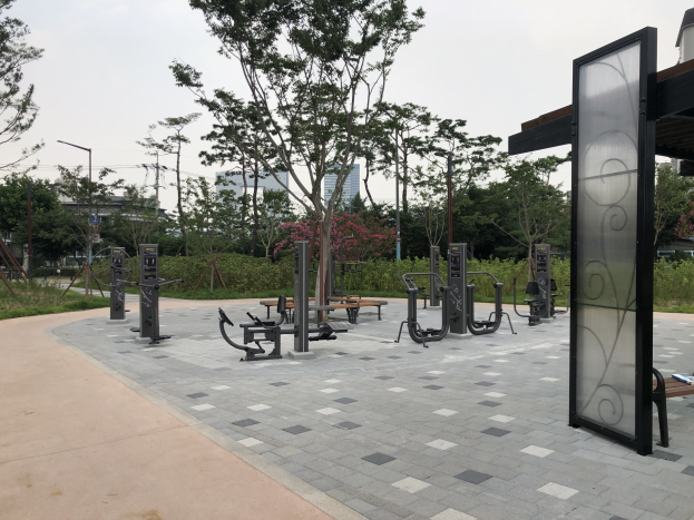 Outdoor park featuring fitness equipment, benches, trees, plants, grass, poles, lights, wires, and surrounding buildings under a sky backdrop.