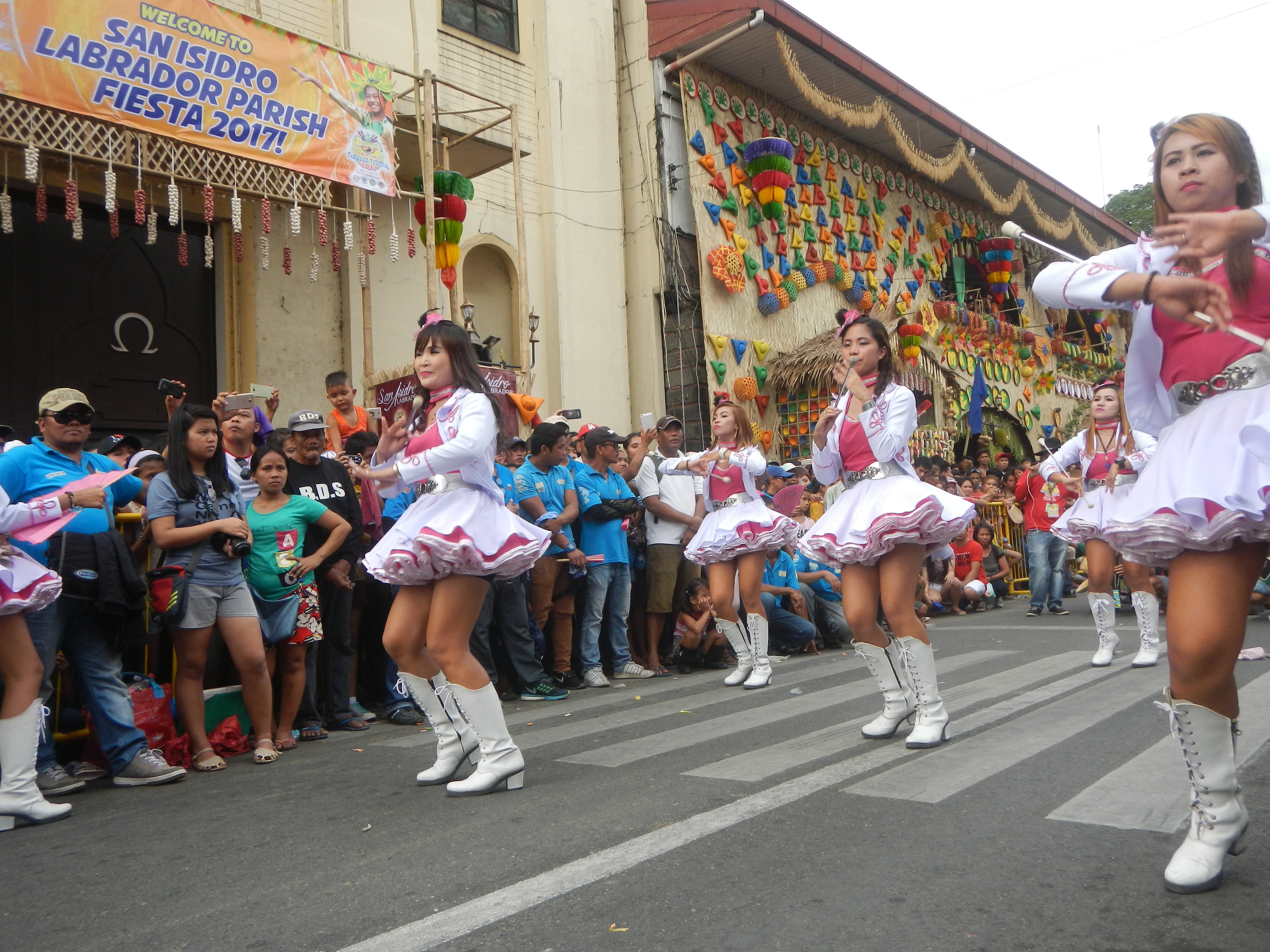 Eine Gruppe von Frauen in pinken und weißen Outfits tanzt auf der Straße, umgeben von einer Menge Menschen, die stehen und zusehen, mit Gebäuden, Bäumen und einem klaren blauen Himmel im Hintergrund.