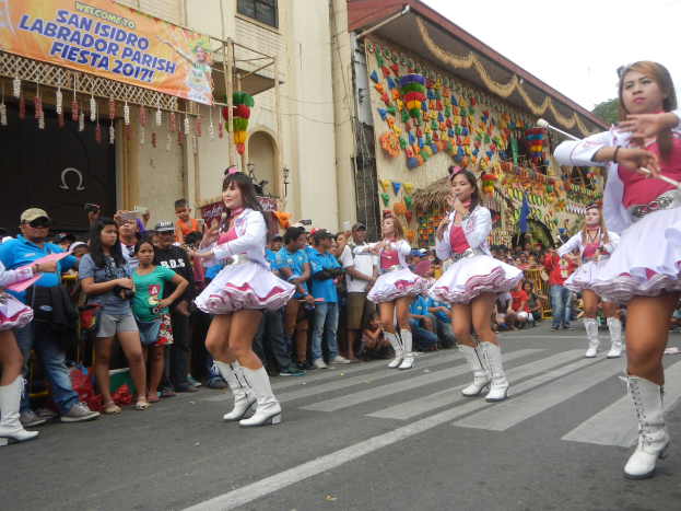 Eine Gruppe von Frauen in pinken und weißen Outfits tanzt auf der Straße, umgeben von einer Menge Menschen, die stehen und zusehen, mit Gebäuden, Bäumen und einem klaren blauen Himmel im Hintergrund.