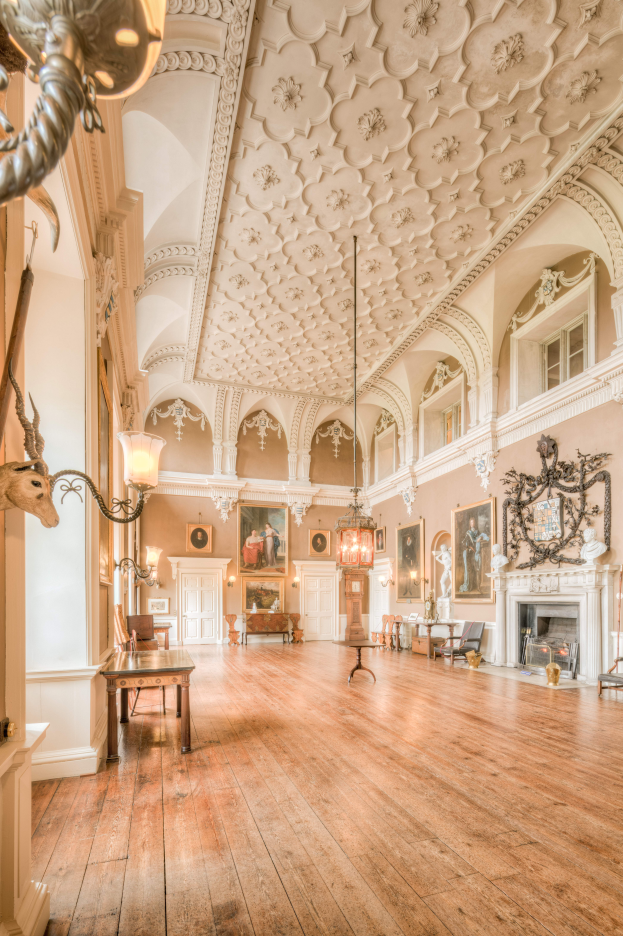 Ein prächtiger Schloss-Saal mit Holzboden, Kronleuchtern, Tischen, Stühlen, einem Kamin, Statuen, Fotorahmen an den Wänden und Fenstern.