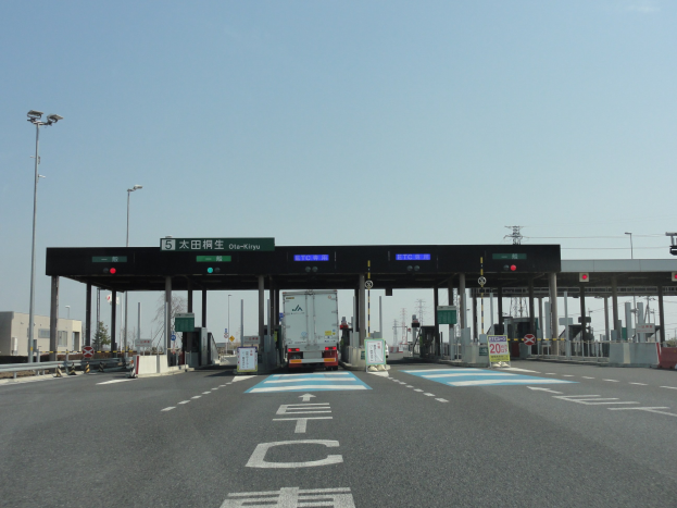Mautstation an der Straße mit einem geparkten Lastwagen, weißen und blauen Markierungen auf der Straße, Laternenpfählen, Schildern, Gebäuden, Bäumen und einem klaren blauen Himmel im Hintergrund.